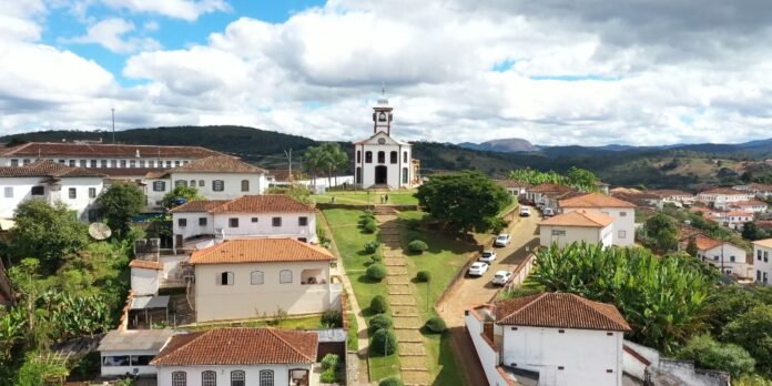 A hillside town with white buildings and red-tiled roofs, a central hilltop church, and a long stairway flanked by trimmed shrubs leading uphill.