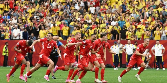 Soccer players in red jerseys celebrate a goal on the field while a yellow-clad crowd cheers in the stands.