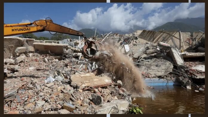 Excavator demolishing rubble at a collapsed-building site with flying dust and debris.