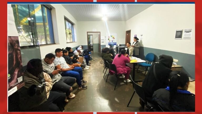 People sit in a waiting room while a woman speaks at a small desk at the front of the room, a banner visible in the background.