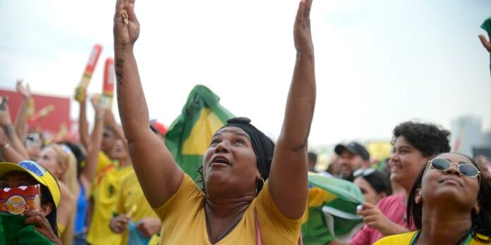 Crowd at an outdoor celebration, several people in yellow shirts; a woman in the center raises both arms toward the sky, with a Brazil flag visible in the background.