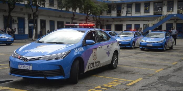 Row of blue police cars with red lights on, parked in a courtyard in front of a building with blue doors and stairs.