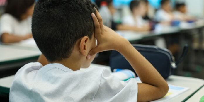 Student seated at a classroom desk with hands resting on the side of the head, back to the camera, classmates blurred in the background (informative pose of concentration or stress).
