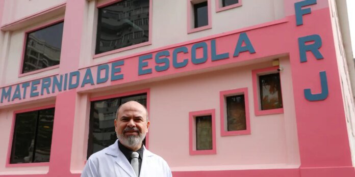 Doctor in a white coat smiling in front of a bright pink maternity school building with blue lettering reading 'Maternidade Escola'.