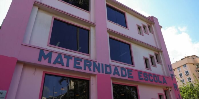 Pink three-story building with large blue-framed windows and a bold sign reading 'Maternidade Escola' along the front façade, modern architectural style.