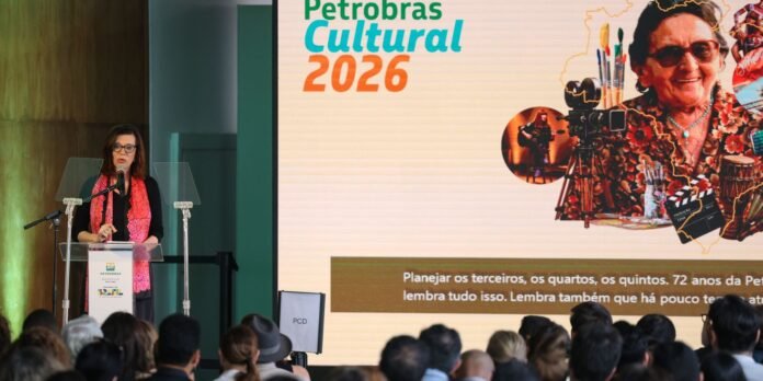 Female presenter with a pink scarf speaks at a podium on a stage, Petrobras Cultural 2026 banner on a large screen behind, with an audience in front.