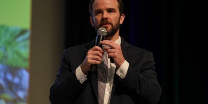 Man in a dark suit speaks into a handheld microphone on stage, with a blurred colorful backdrop on one side.