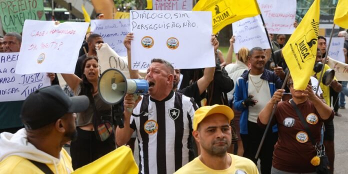 Crowd of protesters on a street, yellow flags, and signs; man in a striped jersey speaks into a megaphone.