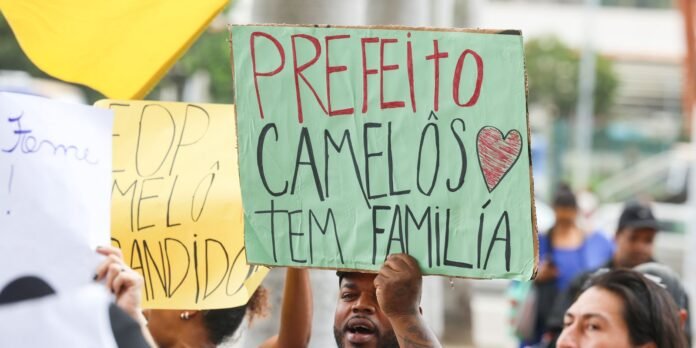 Protest sign reading 'PREFEITO CAMELOS TEM FAMILIA' with a heart, raised at a street demonstration by a man in a crowd.