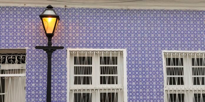 Blue-and-white azulejo tiled wall with a lit black street lamp in front of two white-framed windows with decorative bars.