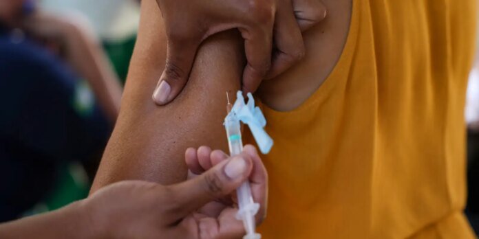 Close-up of a syringe being inserted into the upper arm for vaccination, held by a clinician; the recipient wears an orange top.