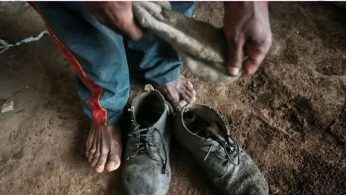 Person kneeling on a dirt floor, handling a dusty boot next to a pair of muddy, worn work boots.