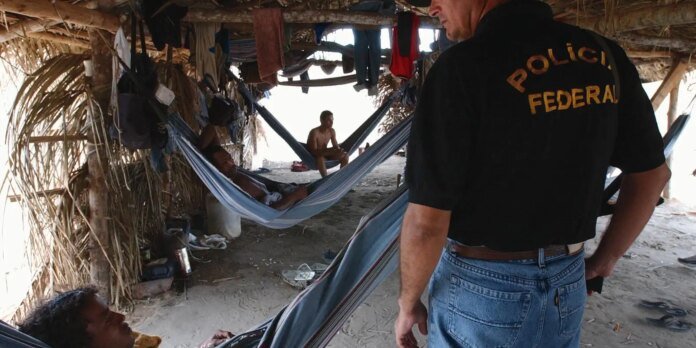 Federal police officer in a black shirt with 'Policia Federal' on back stands beside a beachside hammock shelter where several men rest under a thatched canopy.