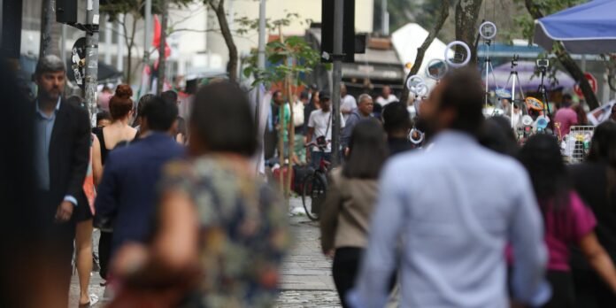 Crowded urban street market with shoppers walking along the sidewalk and vendor stalls visible in the background.