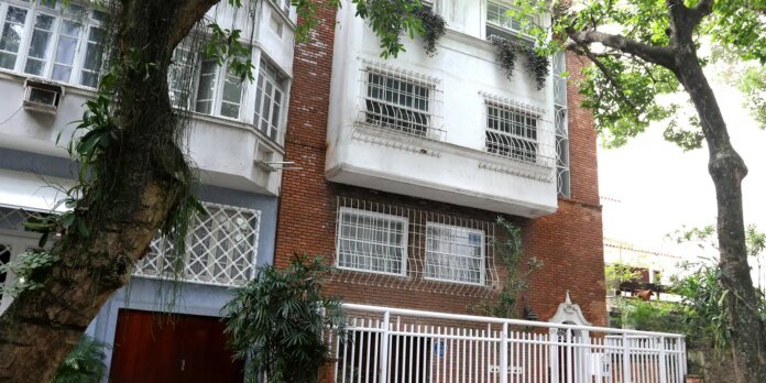 Residential building facade with brick and white walls, barred windows, and a white metal gate, framed by large trees and greenery in front