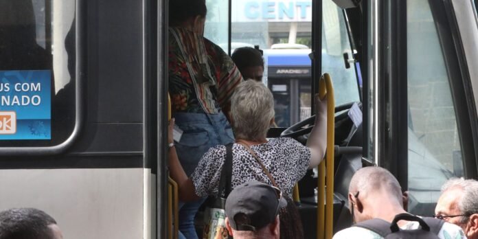 A woman with short gray hair in a leopard-print top stands at the bus doorway, gripping a yellow handrail while boarding a bus filled with passengers.