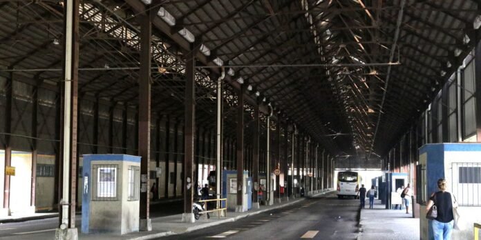 Long covered transit concourse with steel beams, columns, and a bus visible at the far end surrounded by passengers and waiting areas.