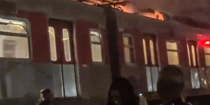 Night scene of a subway train car with flames on the roof and bystanders nearby.