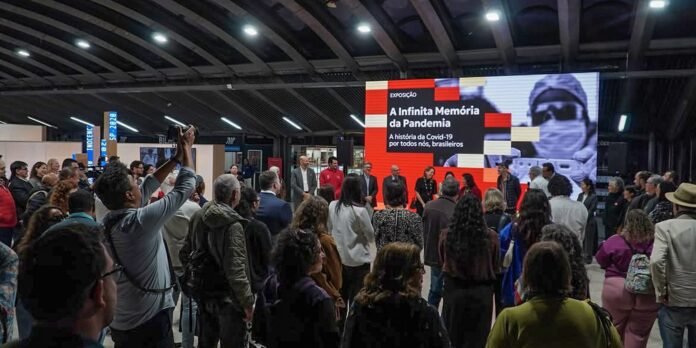 Crowd gathers in an exhibition hall facing a large red-screen presentation about the pandemic memory with speakers at center stage.