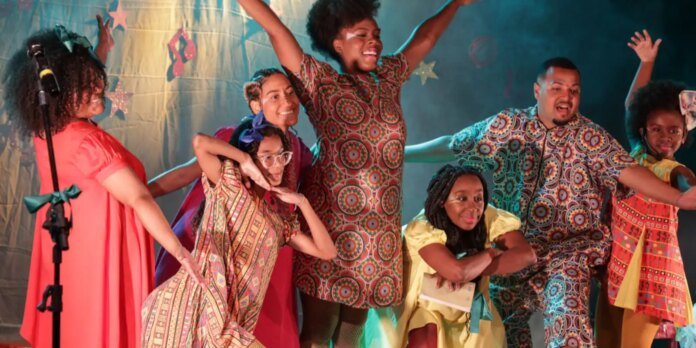 Group of diverse performers dancing on a colorful stage, wearing bright patterned outfits and smiling joyfully.