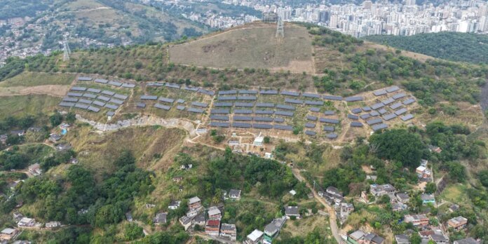 Aerial view of a hillside solar farm with hundreds of panels arranged in rows near a city skyline.