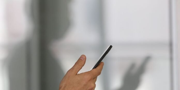 Hand holding a smartphone, with a finger about to tap the screen, in a softly blurred indoor background.