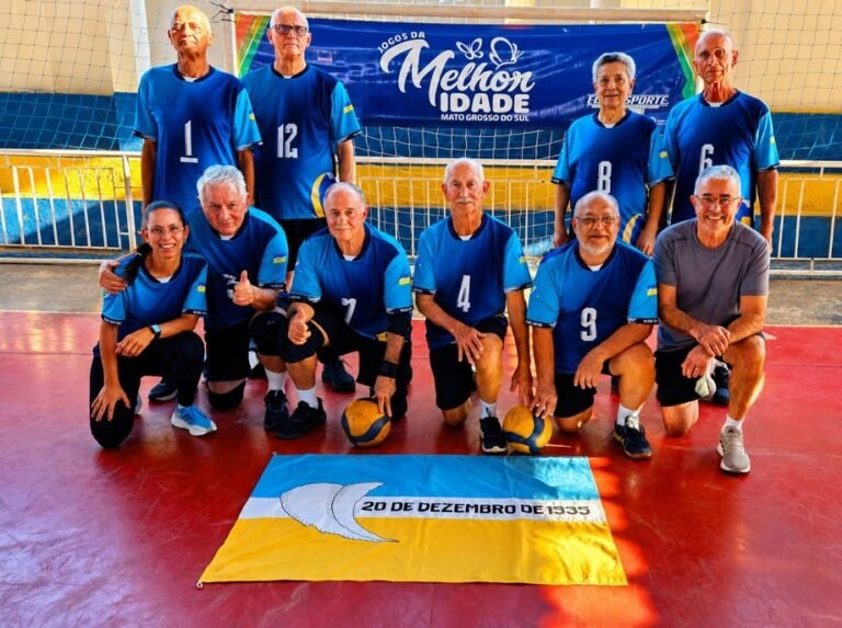 Senior volleyball team of older adults in blue uniforms posing on an indoor court, with a yellow-blue flag on the floor reading '20 DE DEZEMBRO DE 1935' and a banner behind that says 'Melhor Idade'