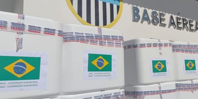 Stacks of white insulated boxes with Brazil flag labels reading 'BRASIL – COOPERAÇÃO HUMANITÁRIA' in front of a 'BASE AÉRE...'(air base) wall area.