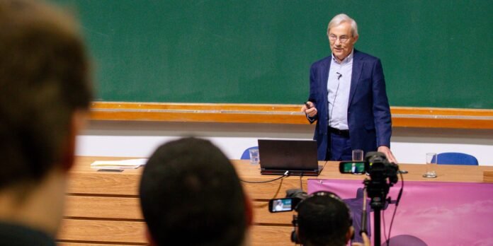 Lecturer in a navy suit stands at a wooden desk with a laptop, addressing an audience while cameras film the lecture in a classroom with a green chalkboard behind him.