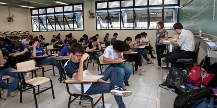 A crowded classroom with students at individual desks taking notes or exams as two instructors supervise at the front near the chalkboard.