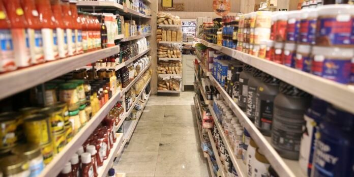 A supermarket aisle with shelves on both sides filled with jars, bottles, and canned goods, leading to the back of the store.