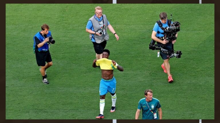 Soccer player in a yellow jersey and blue shorts celebrates on a grass field, lifting his shirt to reveal his abdomen as three cameramen film the moment nearby.