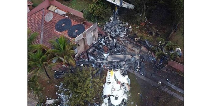 Aerial view of a severely damaged house: collapsed walls, charred debris, and surrounding trees.
