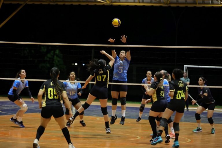 Female volleyball players in blue and black uniforms battle at the net during a night-time indoor match; a blue-team blocker jumps to stop the ball.