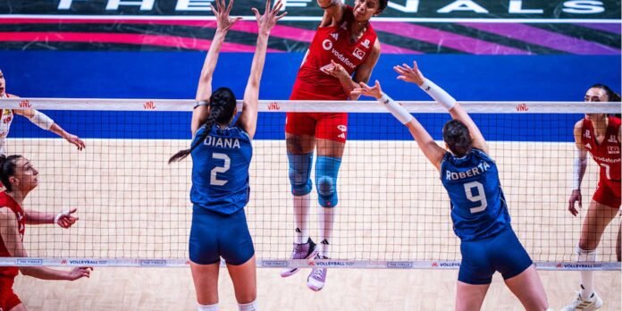 Red-clad volleyball player jumps to spike over blue blockers at the net during a match on an indoor court.] ,0