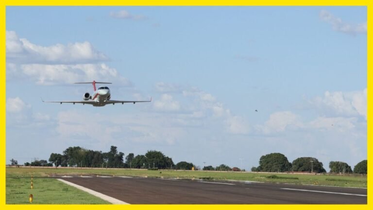 Small private jet lifting off from a runway on a sunny day with green fields and trees in the distance.