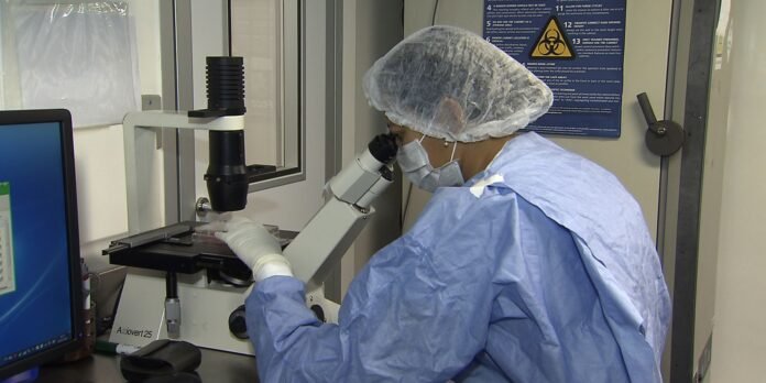 Lab technician in a blue gown, hairnet, and mask using a microscope at a lab bench with a computer nearby.