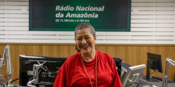 Smiling woman in a red shirt stands in a radio studio with microphones and computers, Rádio Nacional da Amazônia sign visible behind her.