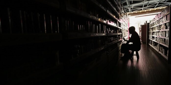 Silhouetted shopper sits on a small stool in a dark store aisle, reaching for items on tall shelves on both sides, light at the end of the aisle.
