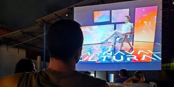 Audience seated in a dark venue watching a large outdoor screen showing a man sitting in a chair on stage with colorful graphics behind him.