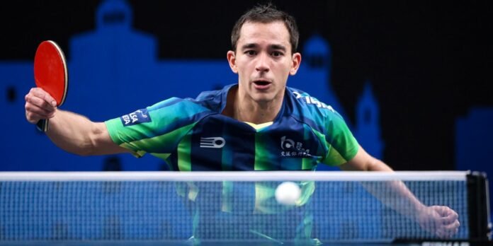 Male table tennis player in a green-blue shirt lunges to hit the ball with a red paddle at a table, net in foreground and blue backdrop behind him.