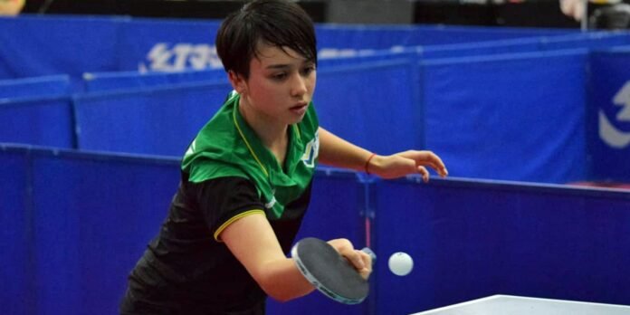 Young female table tennis player in a green and black shirt lunges to hit the ball with a paddle at a blue table backdrop.