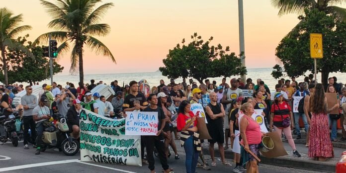 Crowd of protesters on a coastal street at sunset, holding signs demanding workers' rights and beach access.
