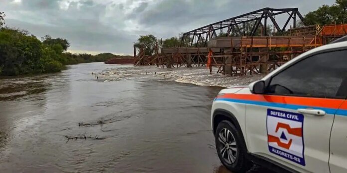 Civil defense vehicle parked by a flooded river with a partially built metal bridge and workers in orange safety gear on scaffolding nearby