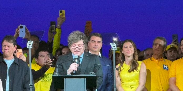 Man in a suit speaks at a podium with a microphone on a stage, a woman in yellow beside him, as supporters in yellow cheer or record with phones behind a blue backdrop.