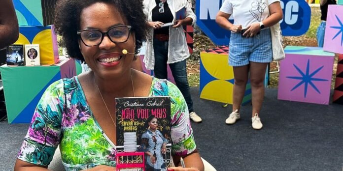 Smiling woman with glasses in a bright floral top sits at a table showing a book titled 'Não Vou Mais' by Cristiane Sobral, with a colorful geometric backdrop and people in the background.