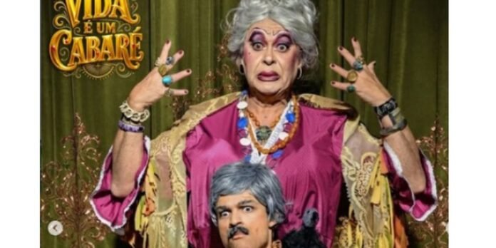 Stage performer in vibrant pink outfit with dramatic makeup and silver wig, posing with hands raised; cabaret banner behind them (Vida é um cabaré).