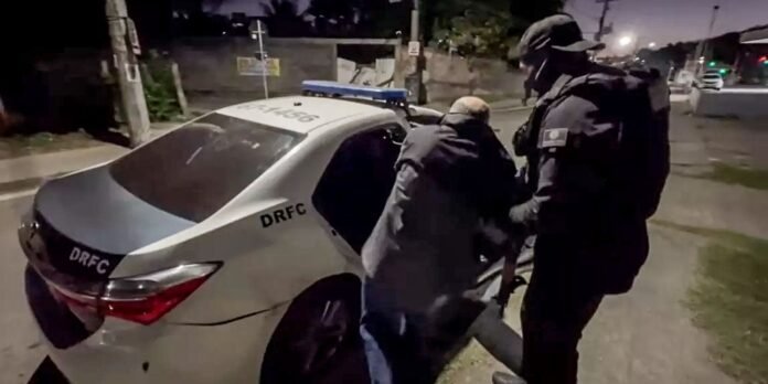 Two officers in tactical gear escort a man toward a police sedan at night; the car bears DRFC markings and a blue light bar on top.