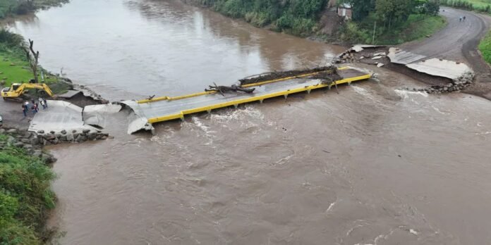 Collapsed concrete bridge over a muddy river with yellow guardrails, wreckage in the middle; construction crew and a yellow excavator assess the site on the left.