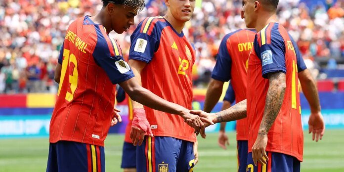 Spain national soccer players in red and blue uniforms shake hands on the field before a match, with a crowded stadium in the background